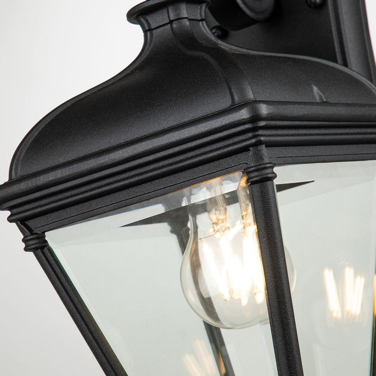 A close-up of the Bayview 1 Light Outdoor Medium Wall Lantern in black reveals its exquisite design featuring ornate detailing and clear glass panels. Inside, a glowing bulb offers warm illumination, enhancing outdoor spaces with its classic and elegant style.