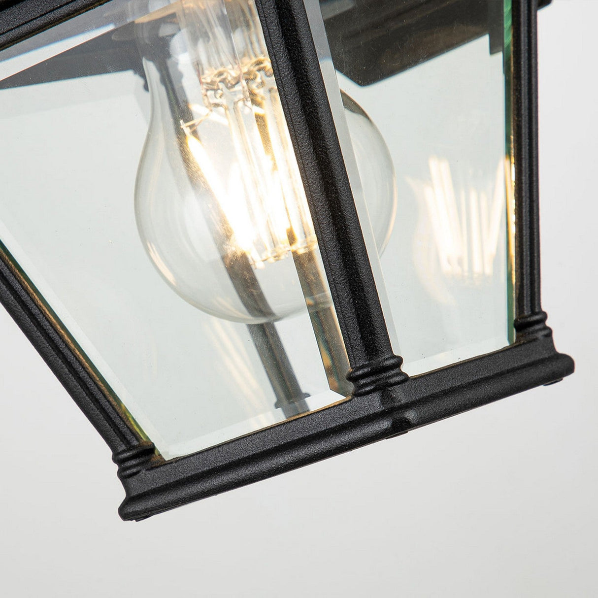 Close-up of the Bayview 1 Light Outdoor Small Wall Lantern - Black, featuring a black frame and glass panels with a glowing bulb inside. Mounted at an angle against a light-colored background, this outdoor lantern offers an IP44 rating for dependable performance in diverse weather conditions.