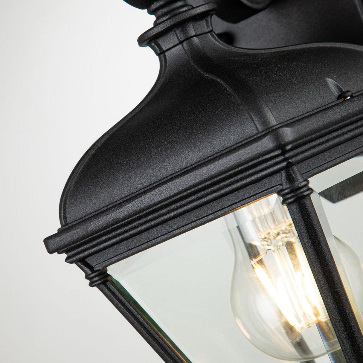 A close-up of the Bayview 1 Light Outdoor Small Wall Lantern - Black showcases its elegant design with glass panels and a glowing filament bulb inside. Perfect for outdoor lighting, this classic piece has an IP44 rating and stands out against a light background.