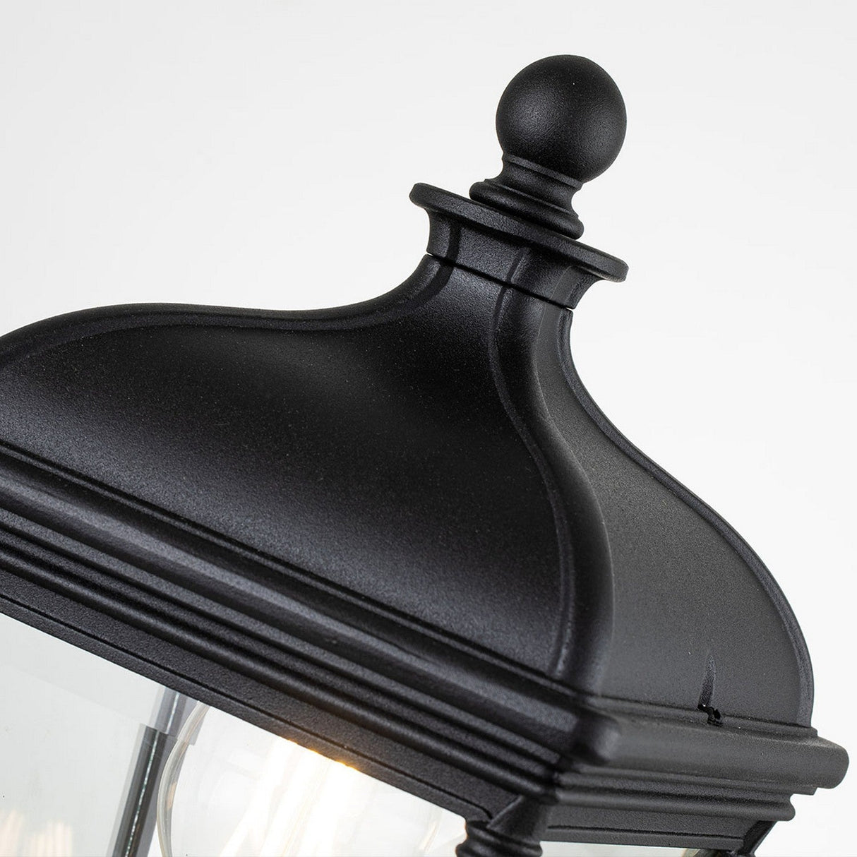 A close-up view of the Bayview 1 Light Medium Pedestal Lantern in black showcases its classical Georgian design, complete with a decorative finial on top. The lantern is framed with clear glass panels that partially reveal the light bulb inside, making it perfect for outdoor lighting. The plain white background highlights its elegance.