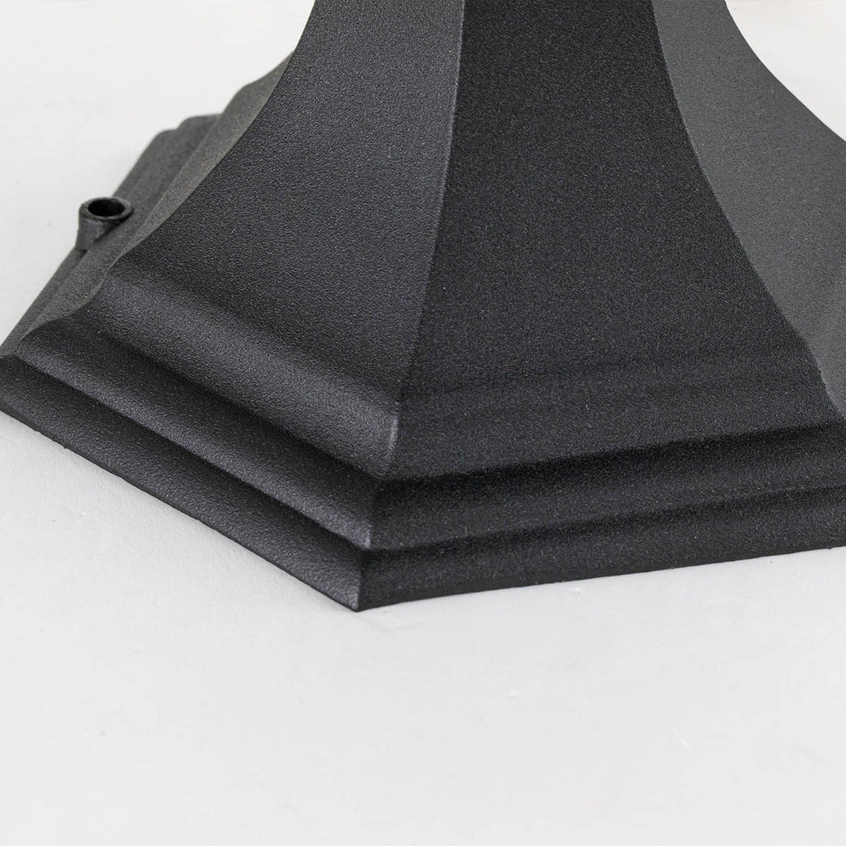 Close-up of the angular and textured base of the Bayview 1 Light Medium Pedestal Lantern - Black, resting on a grey surface. The distinct edges and matte finish impart a hint of classical Georgian design to your outdoor lighting.