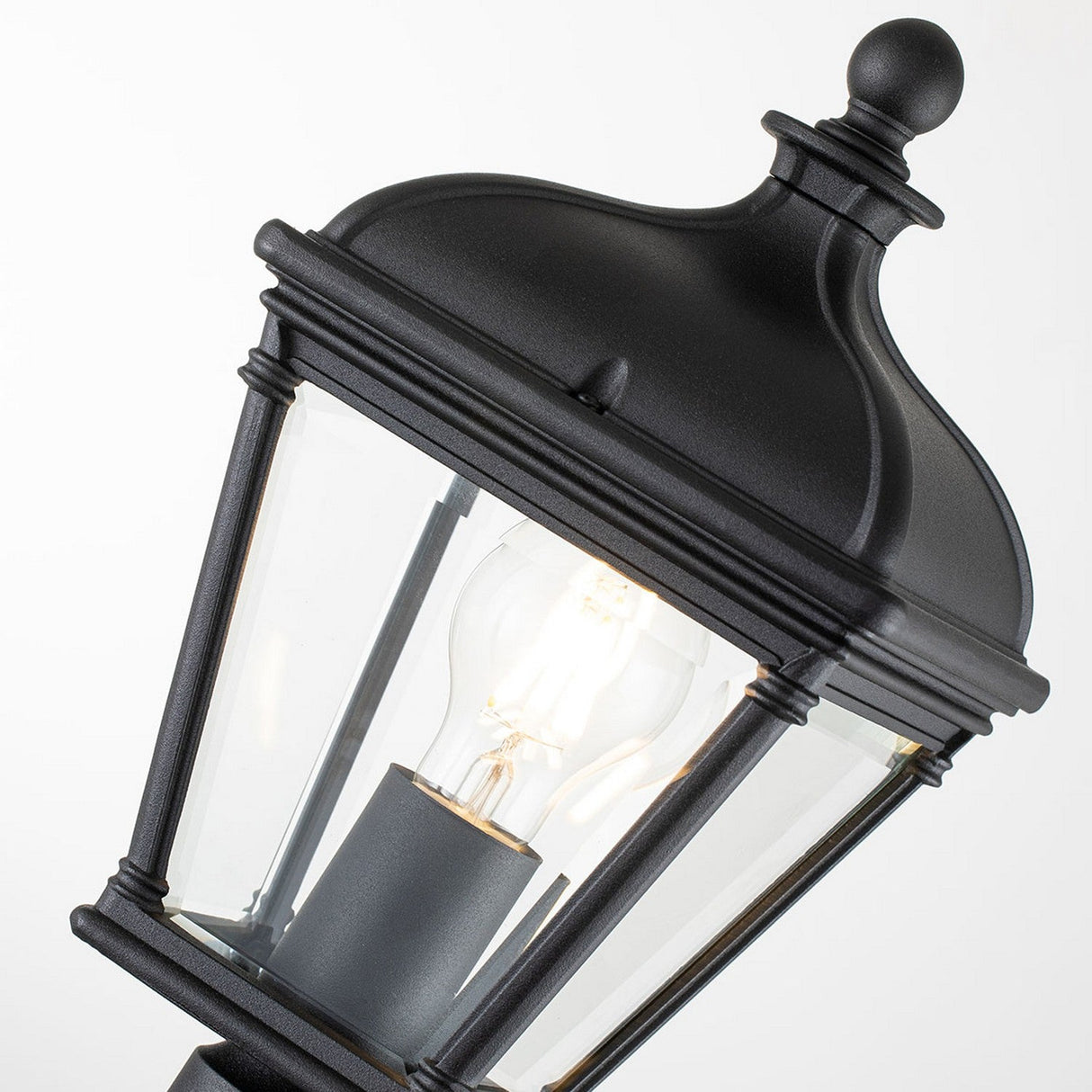 Close-up of the Bayview 1 Light Medium Pillar Lantern in black, featuring a classic design with clear glass panels that reveal a lit bulb inside. This Georgian-inspired piece is slightly tilted against a plain white background, making it an ideal choice for enhancing any outdoor lighting decor.