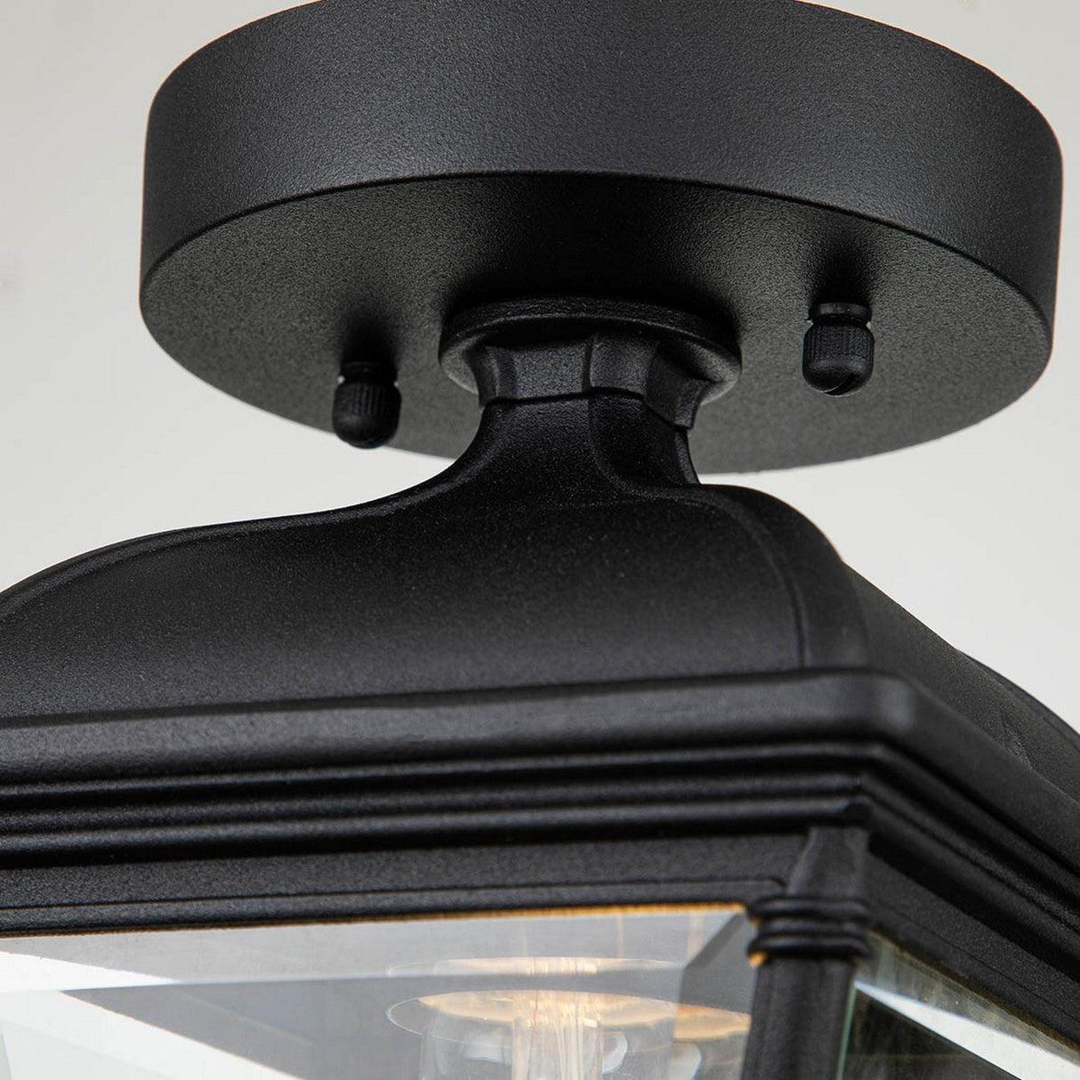 A close-up of the Bayview 1 Light Outdoor Porch Ceiling Light – Black showcases its classic lantern design with clear glass panels. Crafted from a black aluminum frame, this outdoor lighting fixture is mounted on a round base with visible screws and houses a light bulb inside.