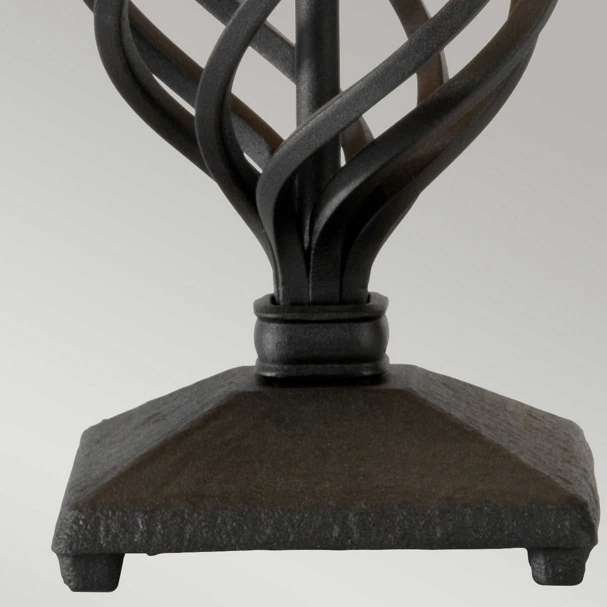 A close-up of the base of a wrought iron table leg showcases artisanal craftsmanship, with twisted metal strips forming a decorative design atop a square base. The dark metal has a smooth finish, echoing the elegant design of the Belfry 1 Light Table Lamp - Black.