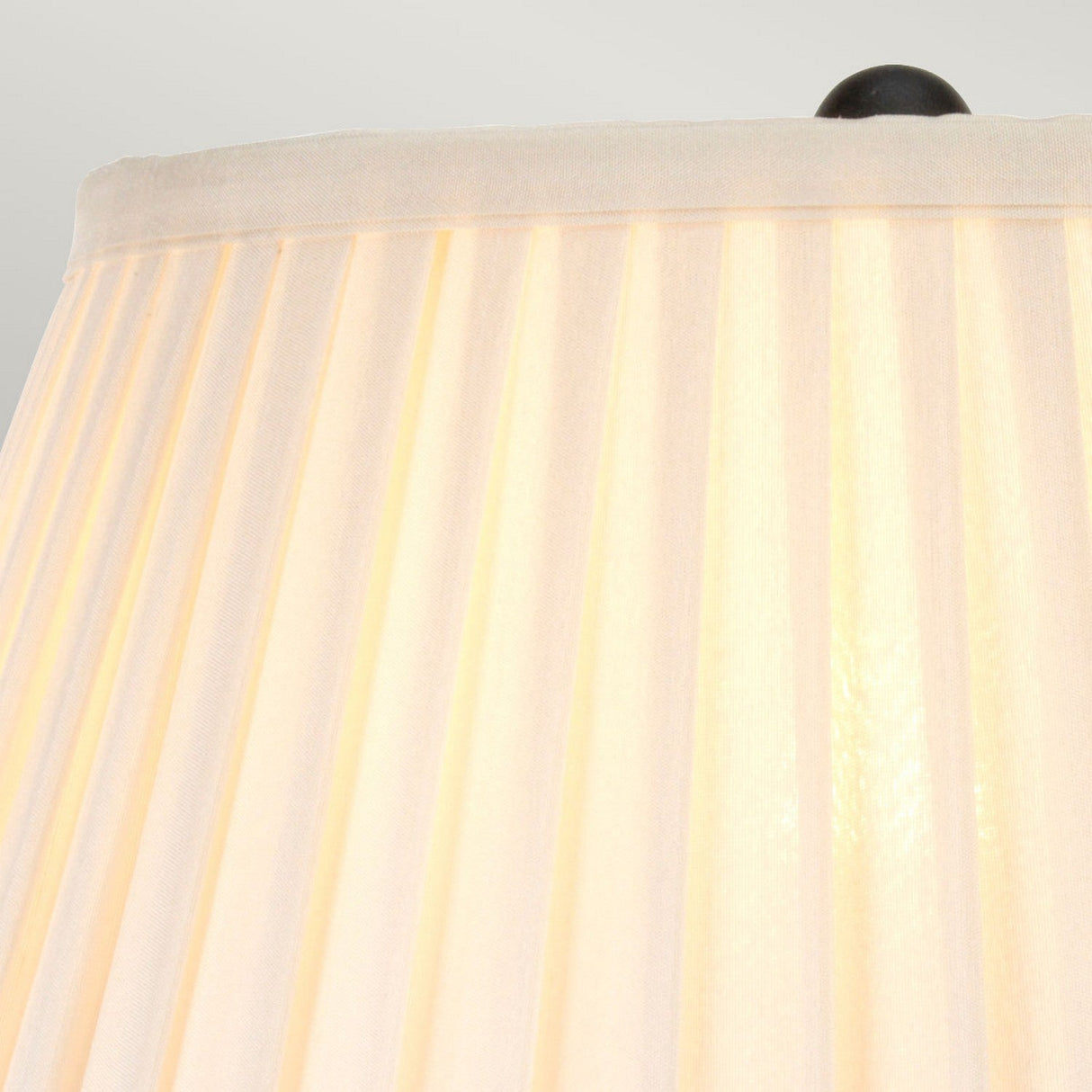 Close-up of the Belfry 1 Light Table Lamp - Black's pleated beige lampshade with a black finial on top, softly illuminated from within. The background is out of focus, emphasizing the elegant design and artisanal craftsmanship of the lampshade.
