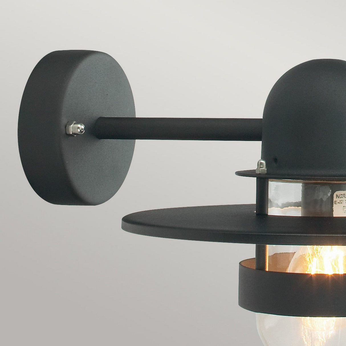 The Bergen Outdoor 1 Light Wall Lantern in black features a modern design with a cylindrical base and round shade, evoking an Art Deco style. The partially visible light bulb is encased in transparent housing, making the lantern an elegant addition to outdoor spaces when installed on a light gray wall.