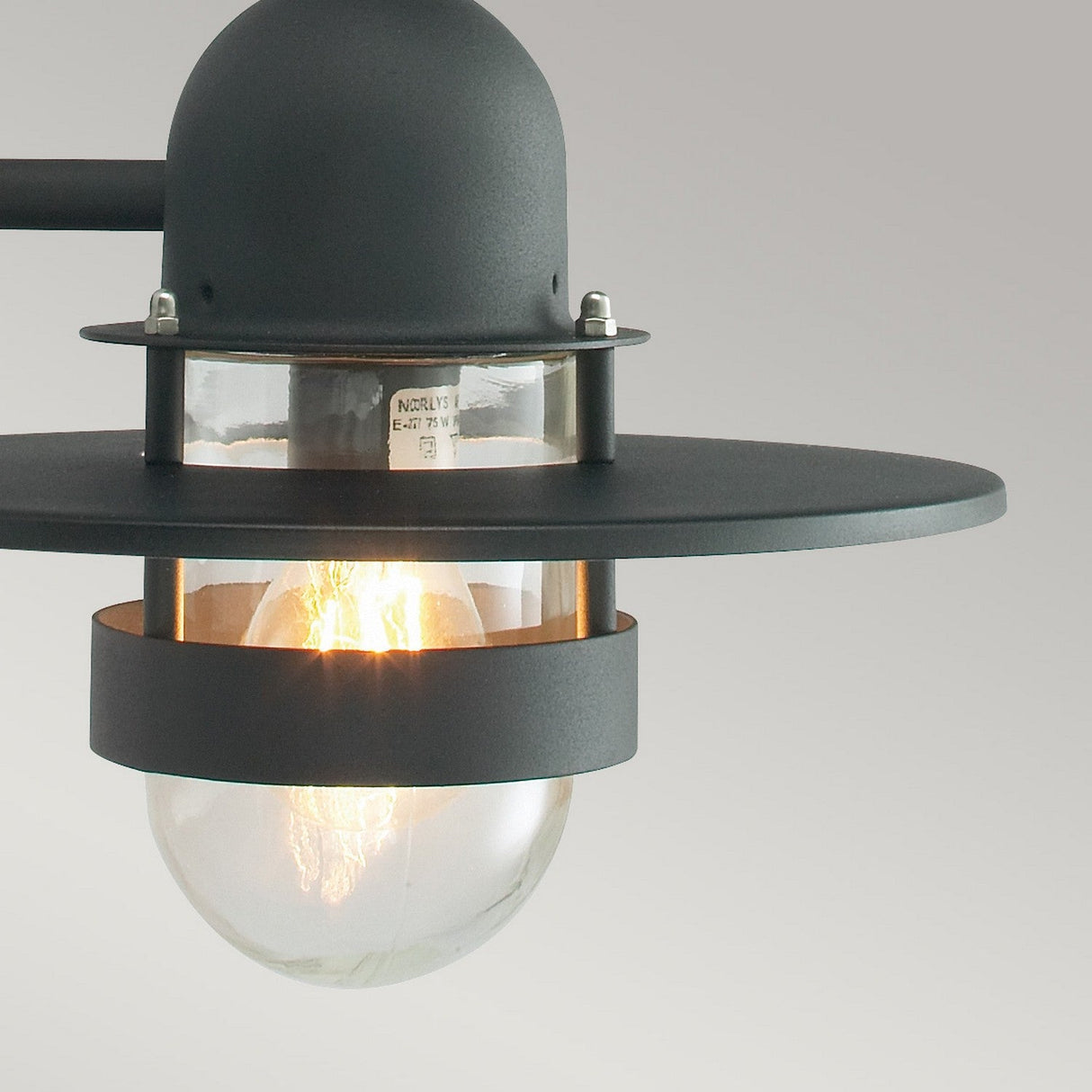 A close-up of the Bergen Outdoor 1 Light Wall Lantern in black, featuring a modern industrial style reminiscent of Art Deco. It has a dark metal frame and a clear glass bulb that casts a warm glow against the light gray background. The lamp's circular top and bottom design make it ideal for outdoor spaces.