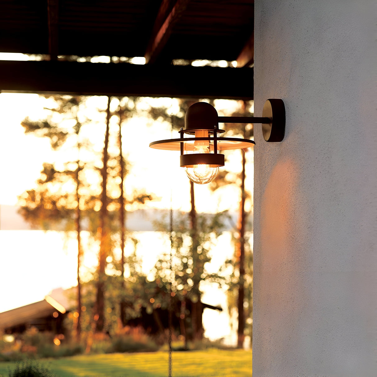 The Bergen Outdoor 1 Light Wall Lantern in black casts a warm glow at sunset, enhancing the serene ambiance of outdoor spaces. The scene unfolds with a view of a tranquil lake and majestic trees, as sunlight filters through, creating a picturesque setting that's both timeless and elegant.