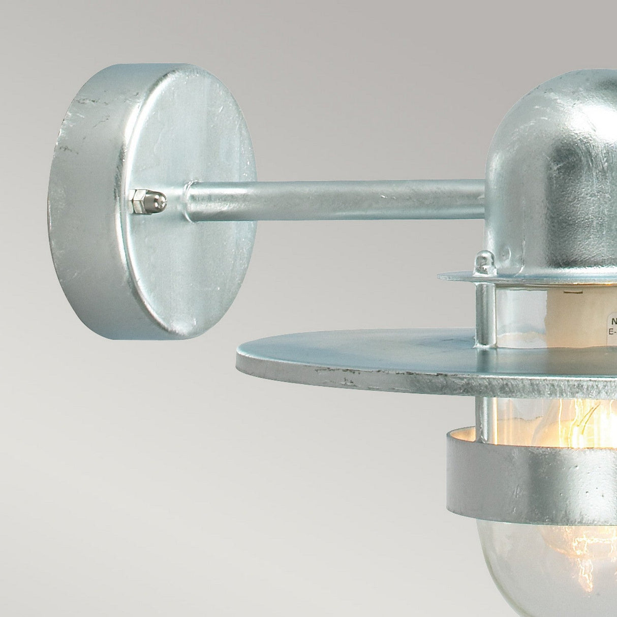 Close-up of the Bergen Outdoor 1 Light Wall Lantern in silver, showcasing its industrial design. The fixture features a round base and a cylindrical rod leading to a flat circular shade, beneath which a clear bulb is partially visible. It has an IP55 rating for enhanced durability.
