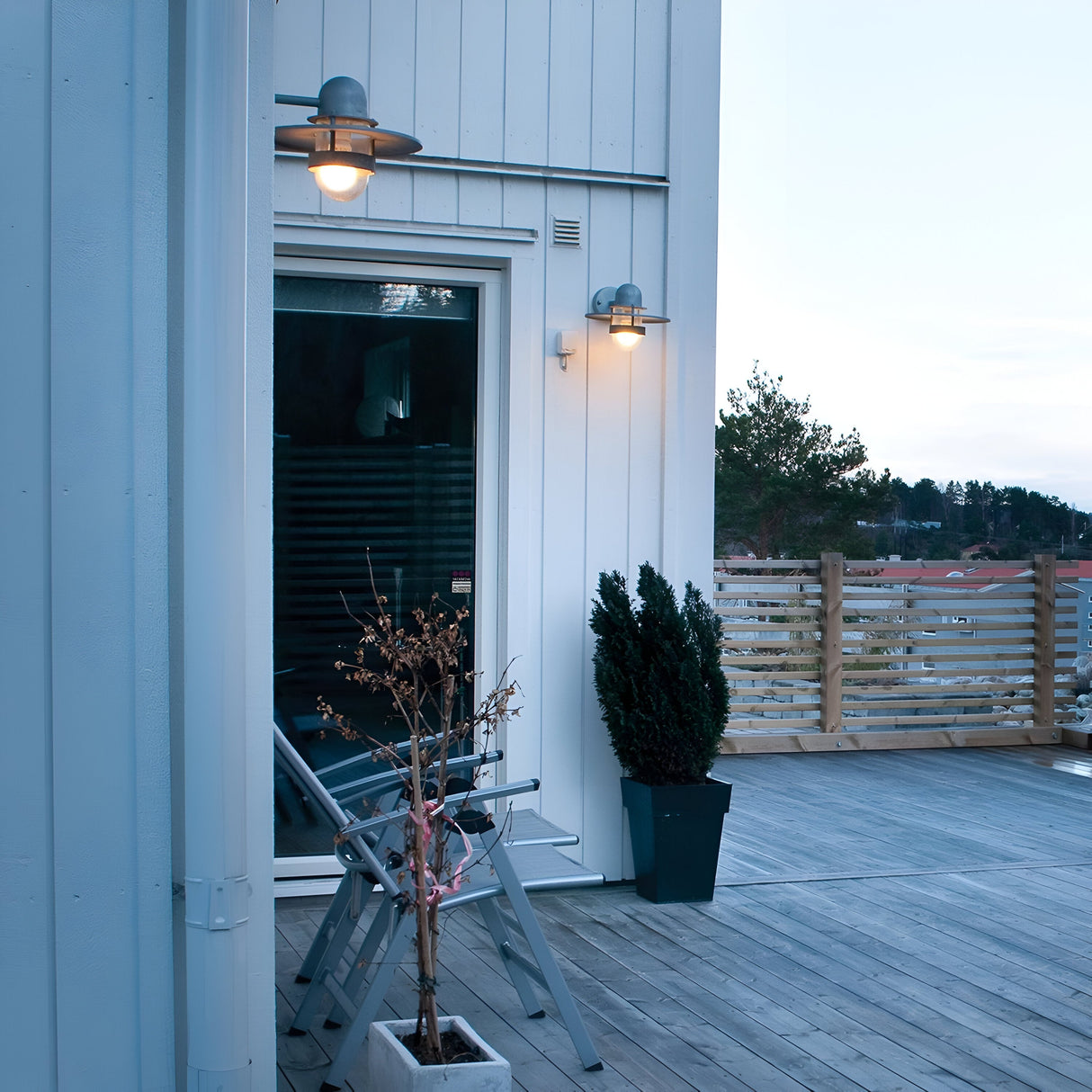 A modern outdoor patio featuring light gray wood decking displays a potted plant and chair set against a white-paneled wall. The area is elegantly illuminated by the Bergen Outdoor 1 Light Wall Lantern in Silver, and is partially enclosed by a wooden fence that provides views of trees and beyond.