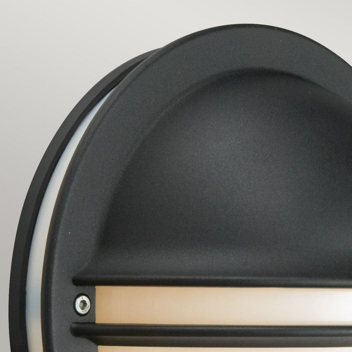 The Berlin Outdoor 1 Light Wall Light - Black is showcased in a close-up shot, featuring a round design with a frosted strip for light diffusion. Its aesthetic appeal is highlighted by two visible screws and a dome-shaped cover. The fixture is set against a plain, light gray background, which accentuates the sleekness of this outdoor lighting solution.