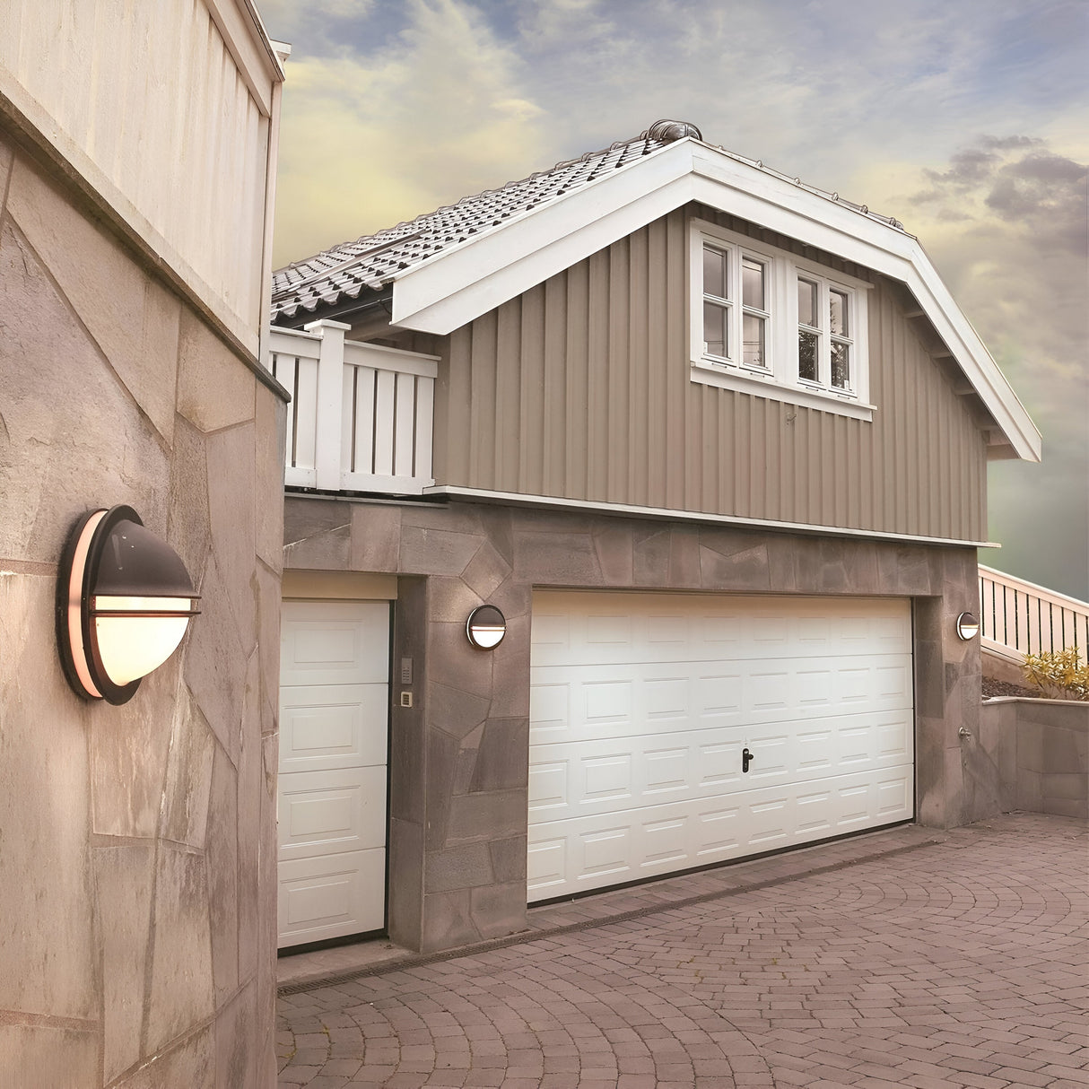 A modern house showcases a double garage with white doors and stone walls. Beside the garage, the Berlin Outdoor 1 Light Wall Light in black adds a sleek touch. The exterior features wooden elements painted gray, with a balcony and windows on the upper level enhancing its design. A cloudy sky offers a moody backdrop to this elegant structure.