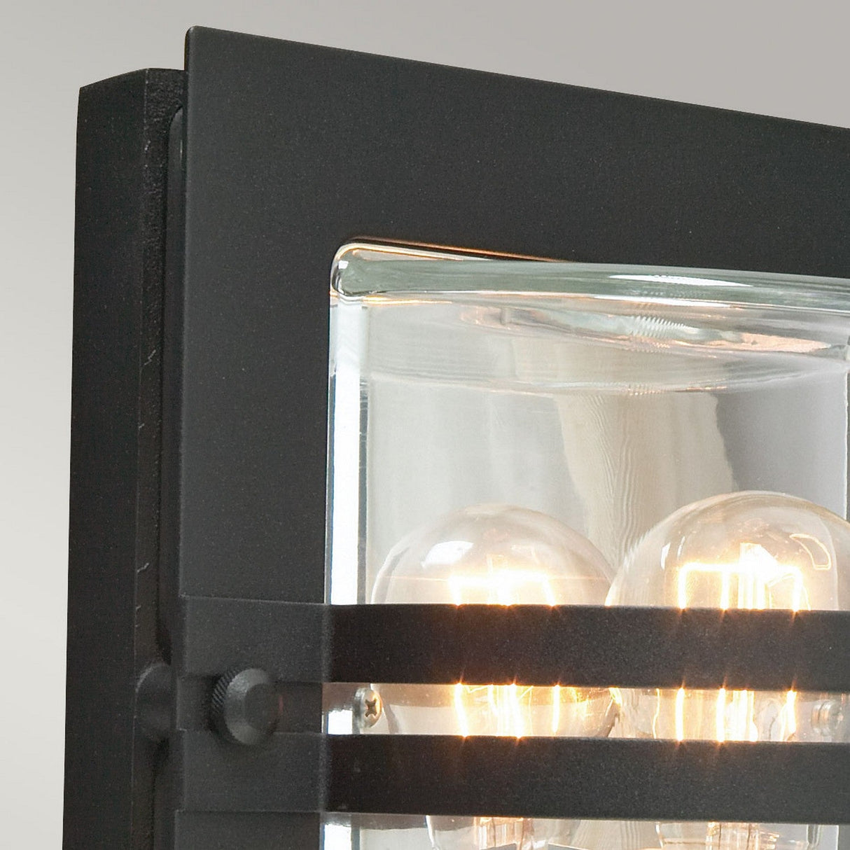 Close-up of the Bern Outdoor 1 Light Wall Lantern in black with clear glass, showcasing visible lit lightbulbs inside. The design features a rectangular frame and sleek horizontal bars, reminiscent of Art Deco lighting. The background is a soft gradient gray.