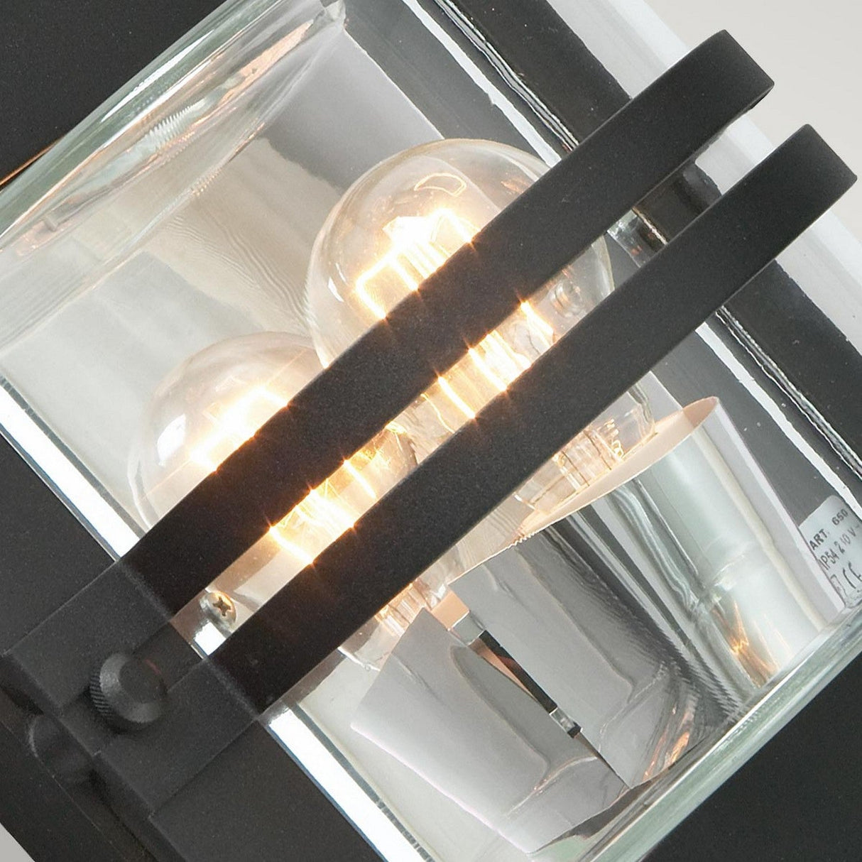 A close-up view of the Bern Outdoor 1 Light Wall Lantern, capturing its modern design reminiscent of Art Deco lighting, showcases a glowing bulb. The metallic frame with black accents and clear glass panels emits a warm glow that creates an inviting ambiance.