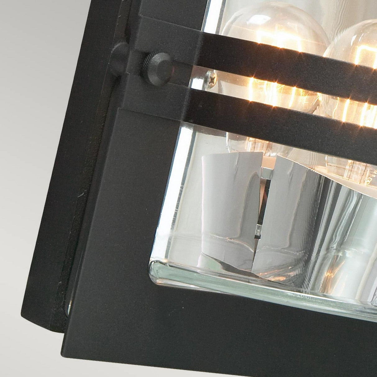 Detailed view of the Bern Outdoor 1 Light Wall Lantern in black with clear glass, featuring its rectangular design with exposed bulbs. The image highlights the texture of the metal frame and reflects inside the glass, evoking an Art Deco style while emphasizing the warm glow of the light.