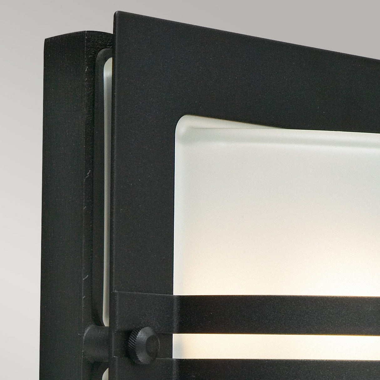 Close-up of the Bern Outdoor 1 Light Wall Lantern in black, showcasing a modern rectangular frame with a frosted glass panel. The sleek design features horizontal bar details inspired by Art Deco lighting and emits a soft, warm glow.