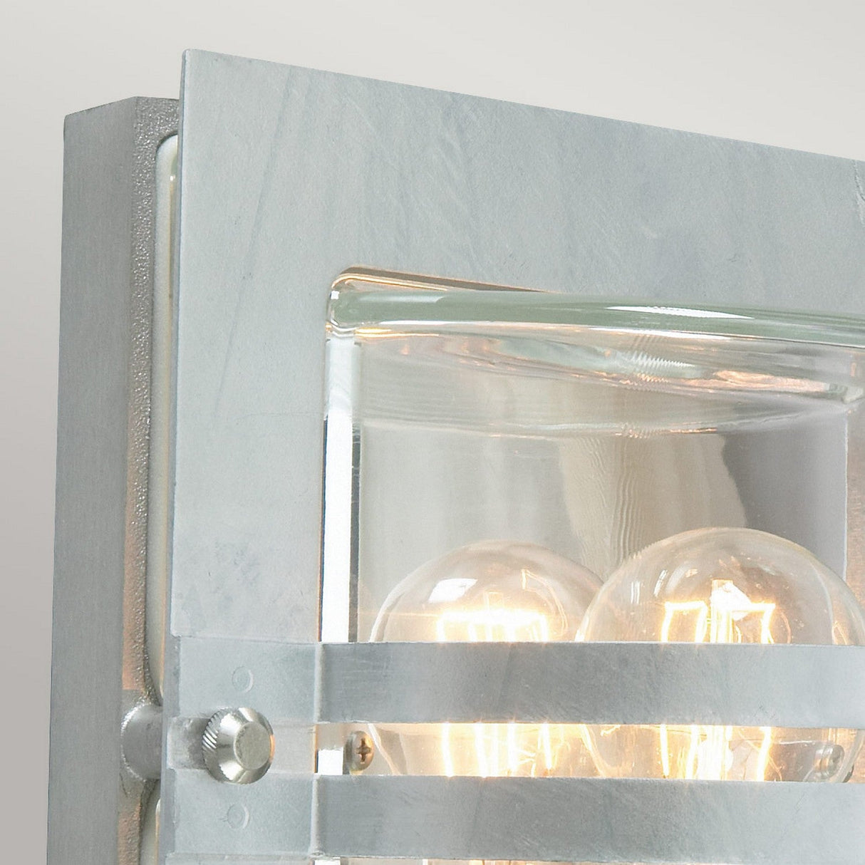 Close-up of the Bern Outdoor 1 Light Wall Lantern, featuring a modern industrial design with two exposed light bulbs housed in a transparent glass and metal frame. Its resemblance to an Art Deco Lantern is enhanced by the brushed silver finish, which beautifully complements the warm glow emitted by the bulbs.