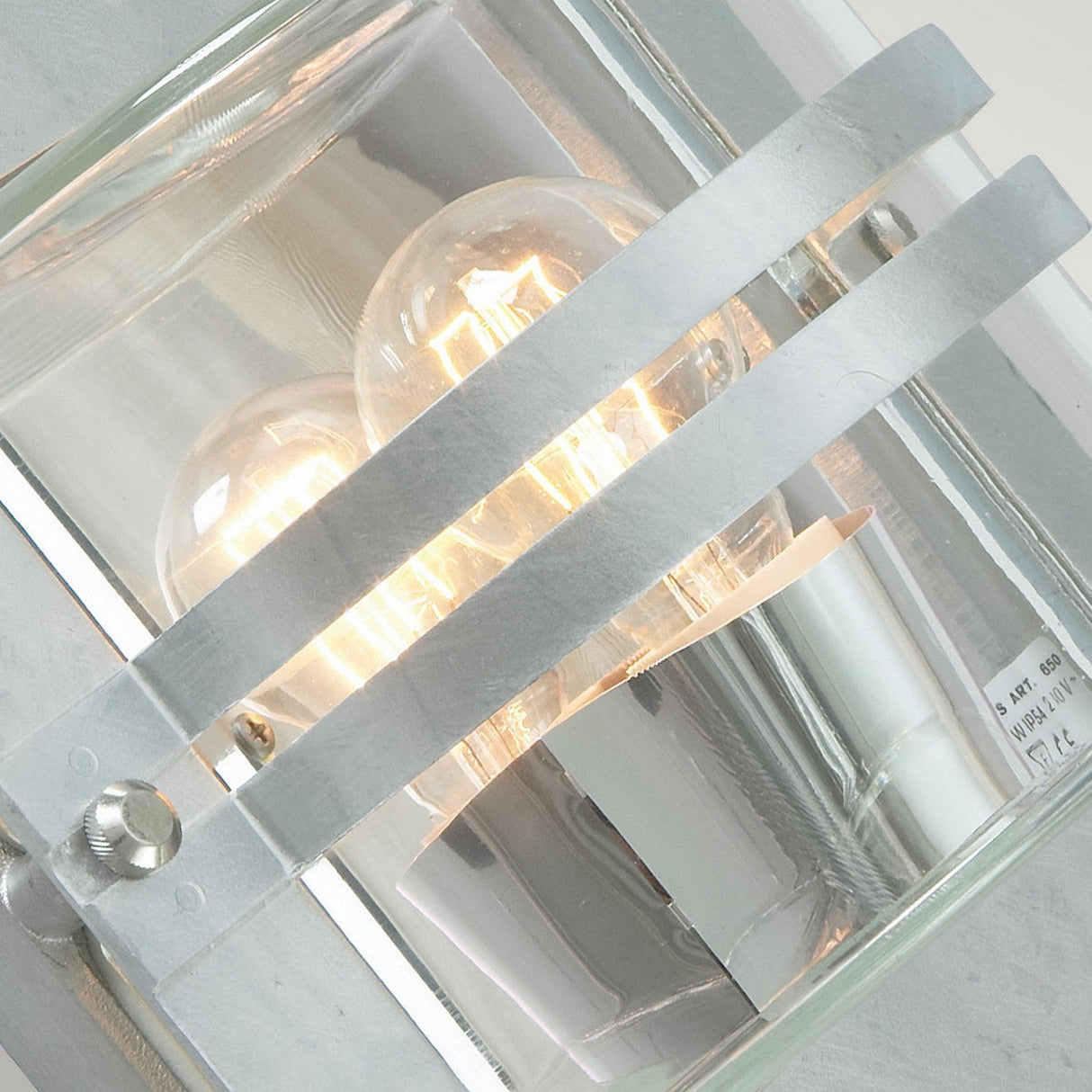 A detailed view of the Bern Outdoor 1 Light Wall Lantern showcases its modern Art Deco design, featuring an illuminated incandescent bulb within a glass and metal housing. Two metal bands cross the front, combining style and durability for outdoor use.