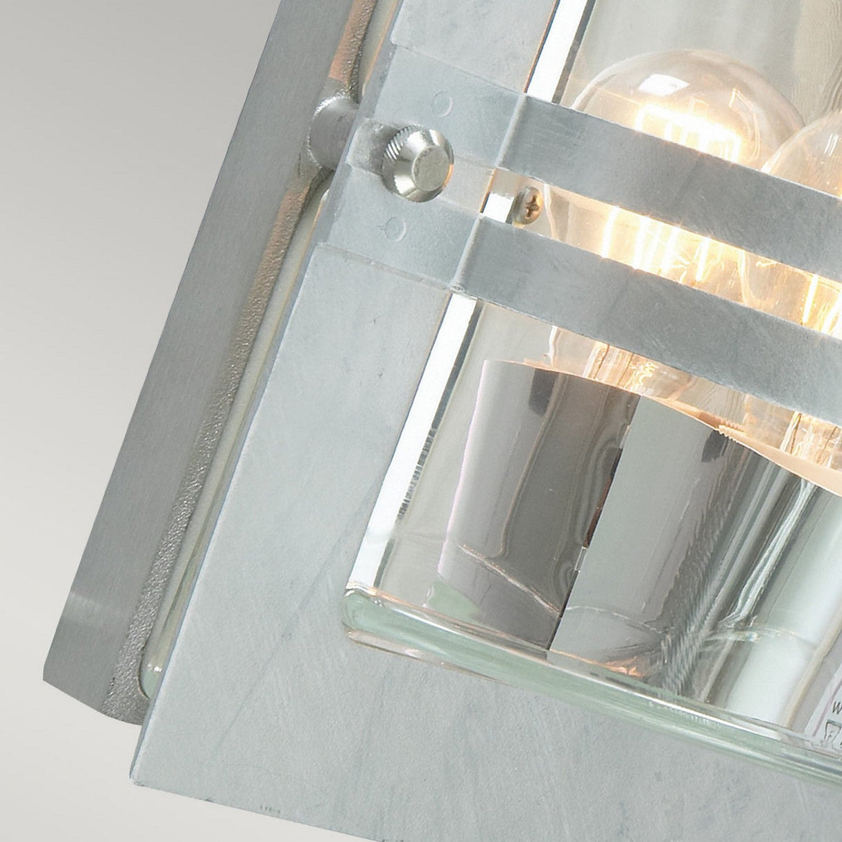 Close-up of the Bern Outdoor 1 Light Wall Lantern in silver, featuring clear glass panels that reveal the glowing filament of a bulb inside. The rectangular frame has a brushed steel finish, captured from a side angle for an elegant effect.