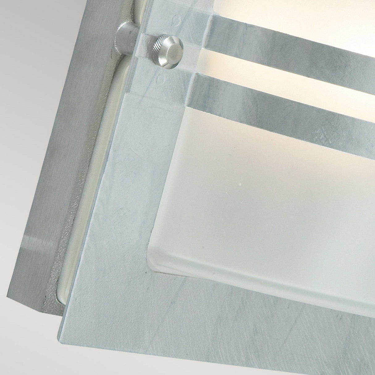 A close-up view of the Bern Outdoor 1 Light Wall Lantern showcases its modern Art Deco style, characterized by a silver metallic frame and frosted glass panel. The design includes horizontal grooves and a screw detail against a neutral background, with softly diffused light enhancing the elegance of outdoor decor.