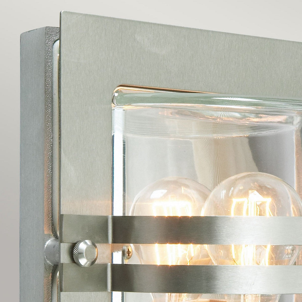 Close-up of the Bern Outdoor 1 Light Wall Lantern, featuring a sleek stainless steel frame and clear glass panels. Inside, one visible light bulb shines brightly, complemented by horizontal metal accents to create a modern and contemporary lantern design.