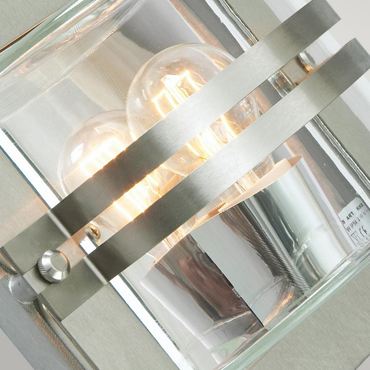 Close-up of the Bern Outdoor 1 Light Wall Lantern in stainless steel, showcasing two exposed incandescent bulbs behind clear glass. The light is on, emitting a warm glow, and the modern design features sleek silver bars across the front with a hint of Art Deco elegance.