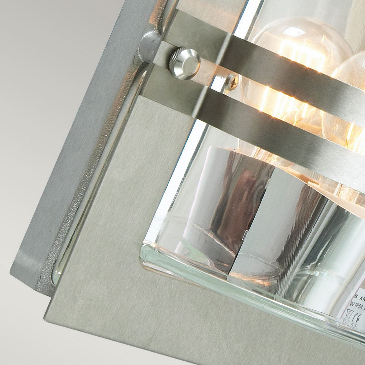 Close-up of the Bern Outdoor 1 Light Wall Lantern, showcasing its transparent clear glass casing and glowing light bulb inside. This Art Deco lighting fixture features stainless steel accents and a neatly aligned design, ideal for modern illumination.