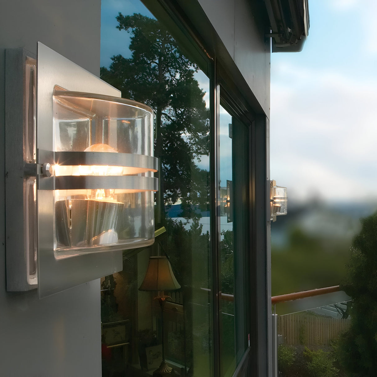 The Bern Outdoor 1 Light Wall Lantern in stainless steel with clear glass illuminates the exterior of the house, its light reflecting off large windows to reveal a lush scene with trees and shimmering water, enhancing the sleek elegance of this modern lantern.