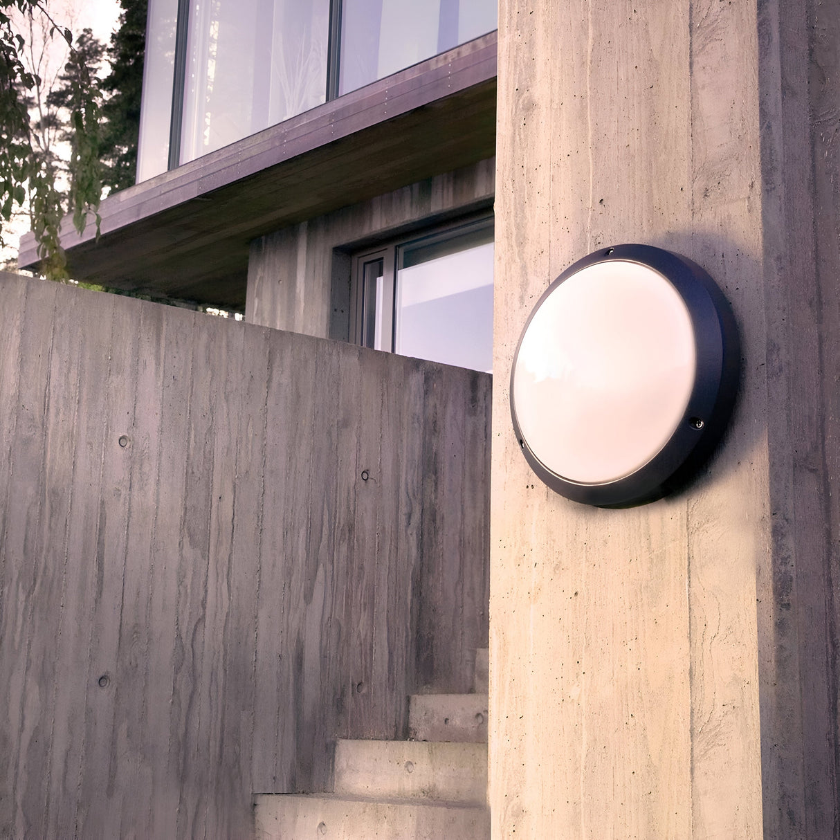 The Bornholm Outdoor LED Wall Light in graphite is installed on a textured concrete wall of a contemporary building. This IP65-rated fixture enhances the structure's sleek lines and expansive windows, with a staircase leading to an upper level. Lush trees can be seen in the background.