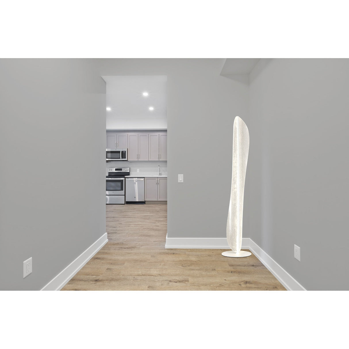 A modern kitchen with gray cabinets and stainless appliances is seen through a doorway. In the foreground, the Bianca LED Floor Lamp 30W 3000K - White stands tall against a light gray wall on hardwood flooring.
