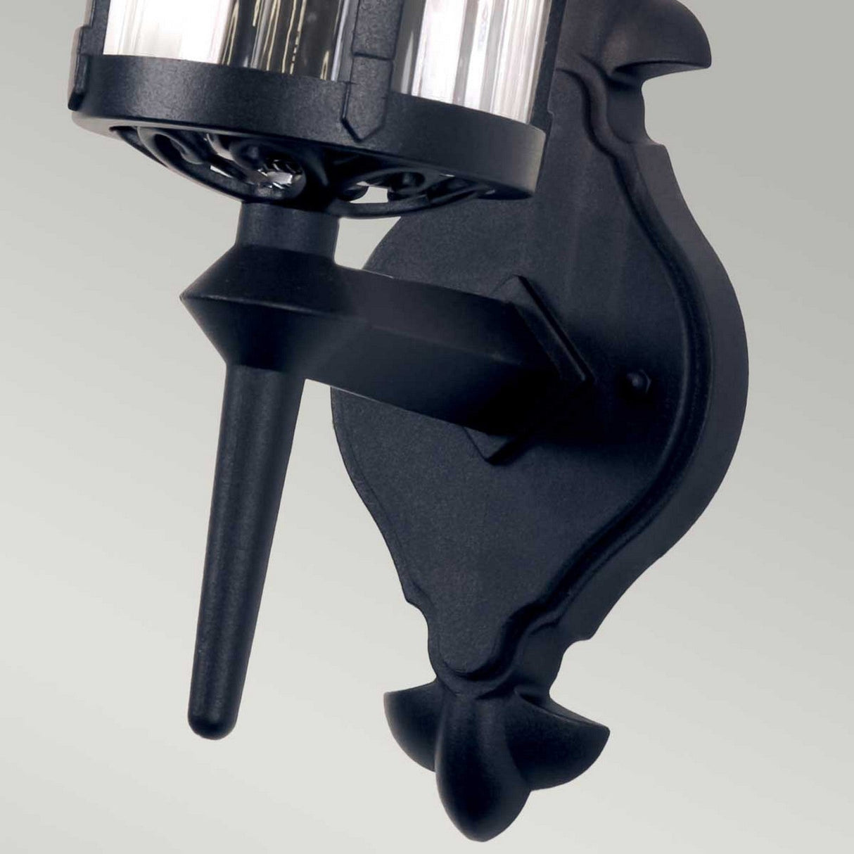 Close-up of the Hereford 1 Light Outdoor Wall Lantern in black, featuring a decorative base and cylindrical glass covering. This wall-mounted light is crafted from die-cast aluminum and is displayed against a plain light-gray background.