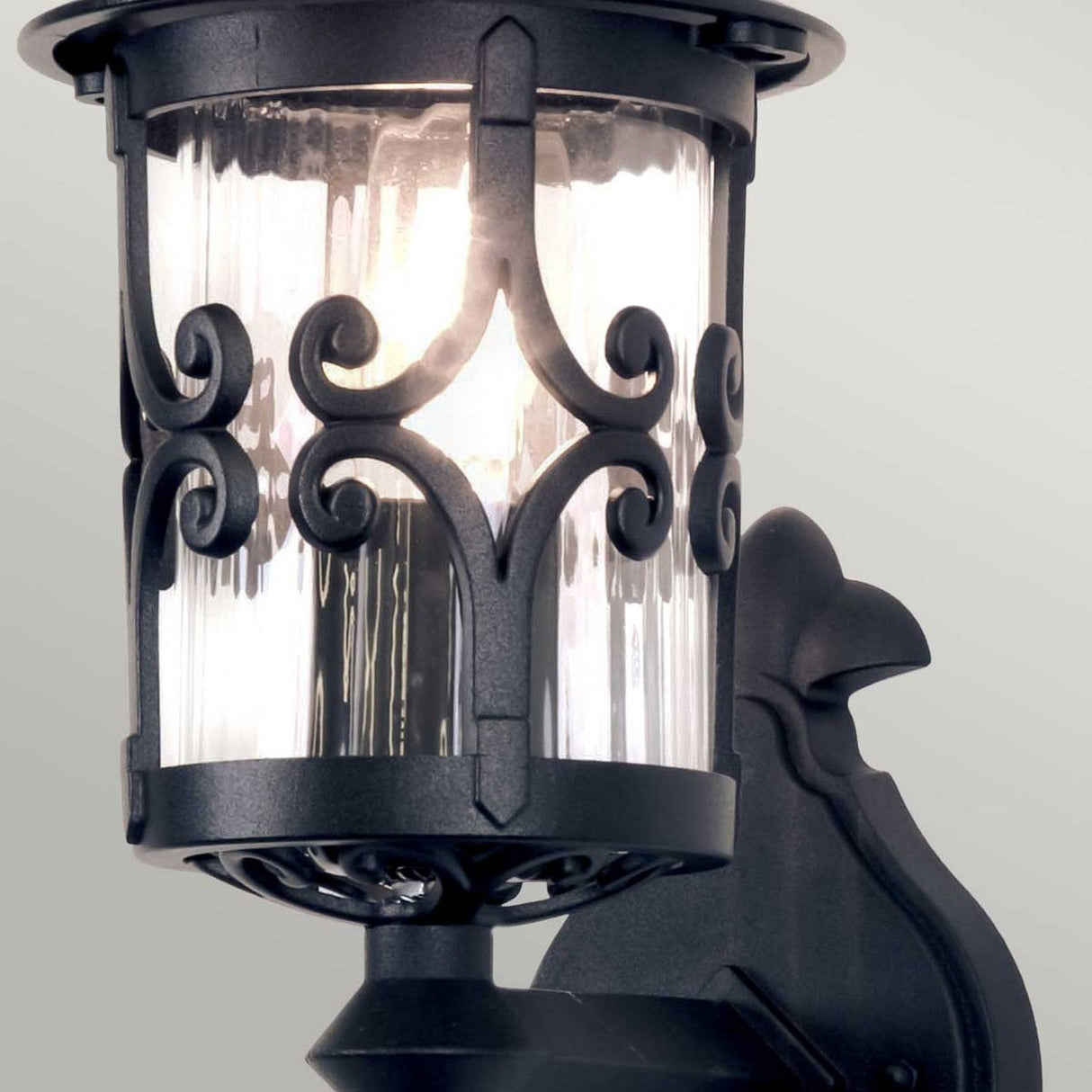 The Hereford 1 Light Outdoor Wall Lantern in black showcases a lantern-style design with intricate scrollwork crafted from die-cast aluminum, surrounding a clear glass casing that encloses the bulb. It is set against a simple, light gray backdrop.