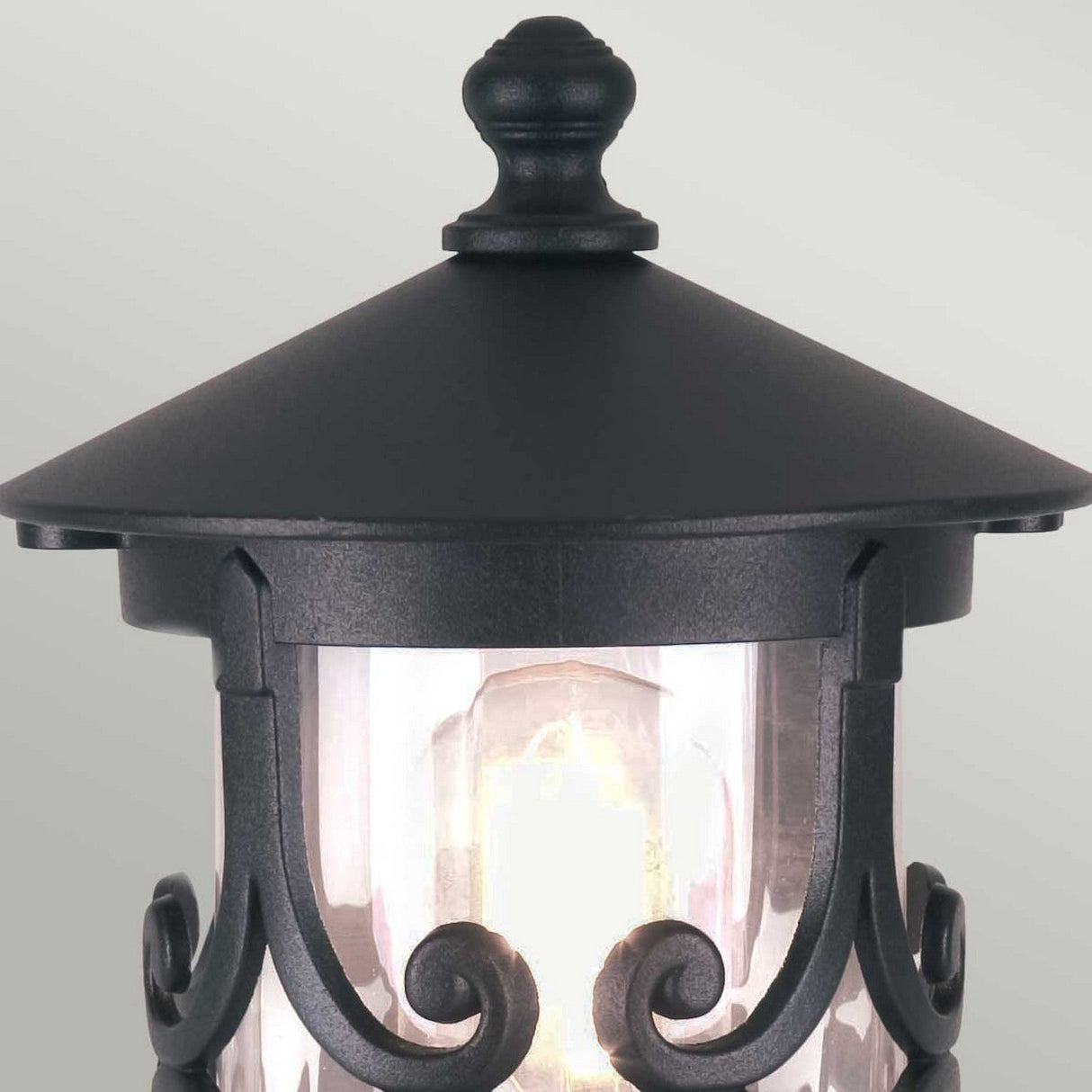 The Hereford 1 Light Pedestal Lantern - Black showcases a black lantern-style with intricate scrollwork, complete with a clear glass casing that reveals the bulb inside, all set against a light gray background.