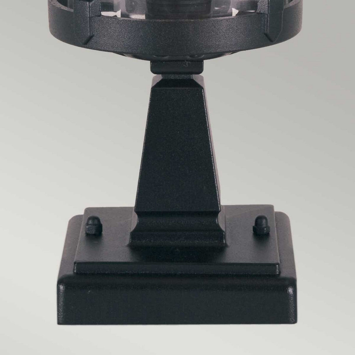 Close-up of the Hereford 1 Light Pedestal Lantern in black, featuring a square base and geometric column. It appears to be a stand, possibly for holding an object or display case. The background is a neutral gray.