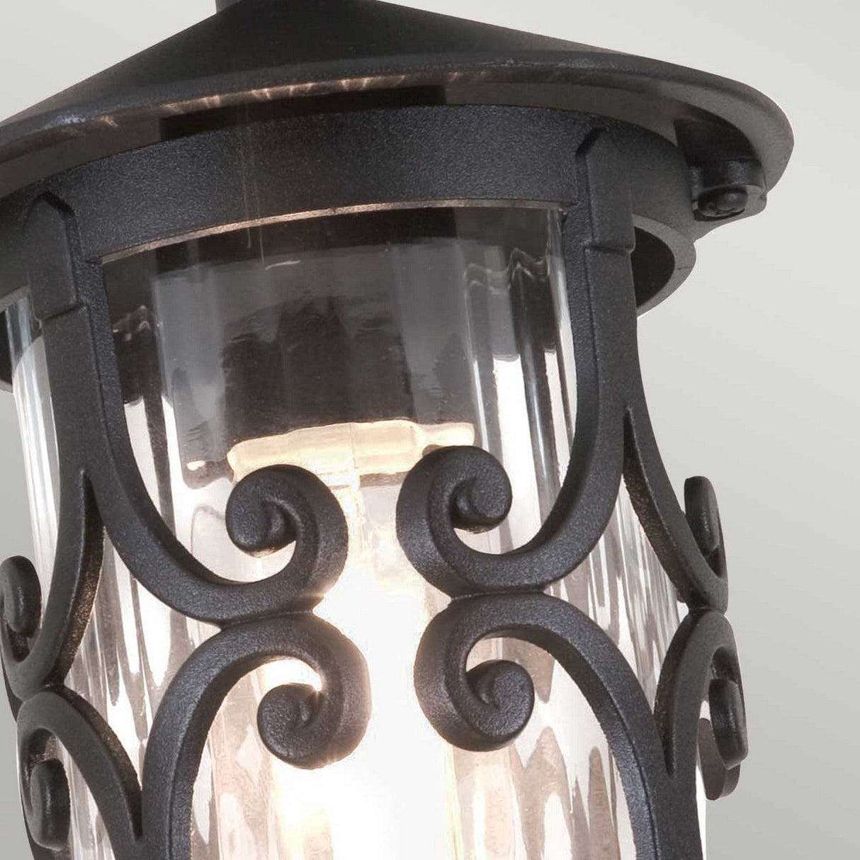 A closer look at the Hereford-1 1 Light Outdoor Porch Lantern - Black reveals a traditional lantern design with elegant black metal scrollwork enveloping a glass enclosure. The warm glow from within highlights the intricate craftsmanship, beautifully contrasting against a plain backdrop to showcase this sophisticated outdoor fixture.