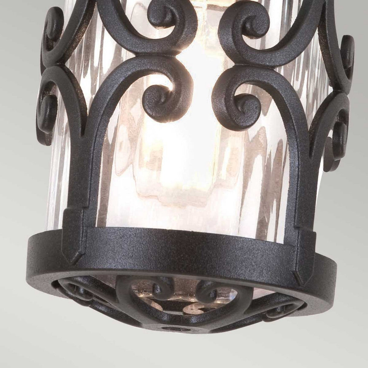 A close-up of the Hereford-1 1 Light Outdoor Porch Lantern shows its elegant black wrought iron scrollwork, reminiscent of a traditional lantern design. The ornate framework encases a clear glass cylinder, revealing the glowing bulb within. The porch light is set against a neutral light gray background.