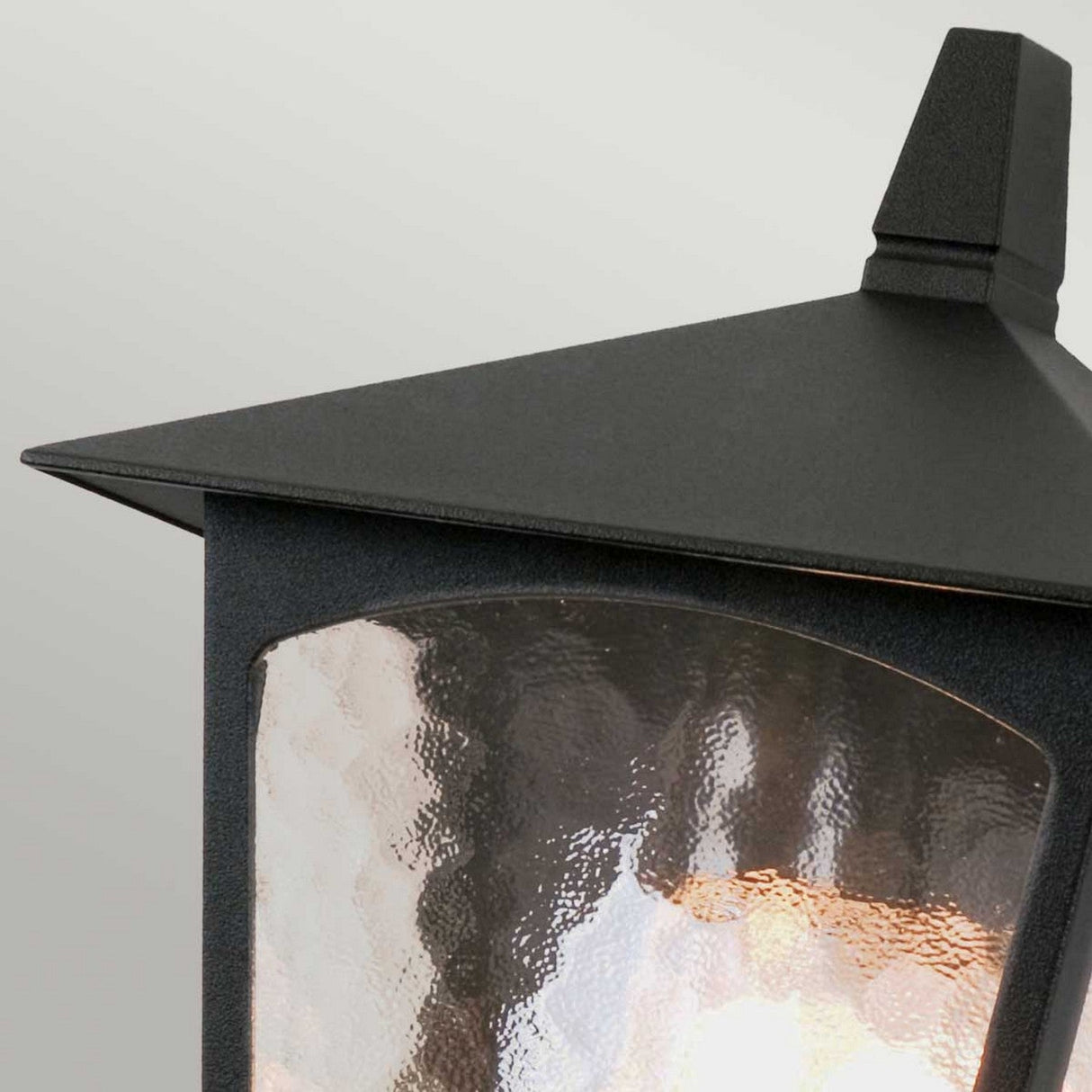 A close-up of the York 1 Light Outdoor Flush Light in black, featuring a modern design crafted from die-cast aluminum and highlighted by a textured glass panel, set against a light gray background. The geometric top and sides of the lantern enhance its contemporary aesthetic.