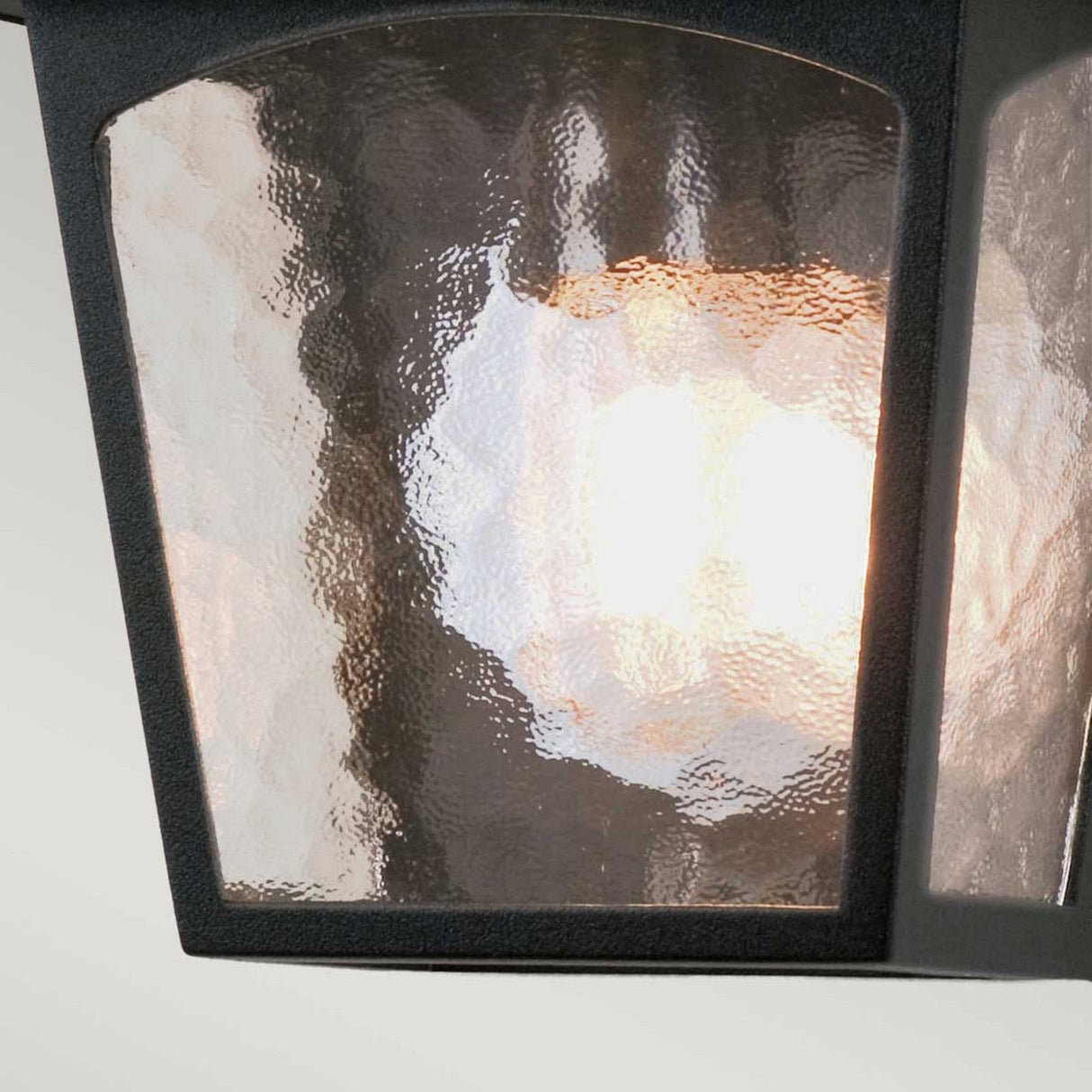 Close-up of a glowing bulb within the textured glass of the York 1 Light Outdoor Flush Light - Black, surrounded by a black metal frame. This fixture is crafted from die-cast aluminum, offering a soft and diffused light pattern that elegantly complements traditional lighting styles.