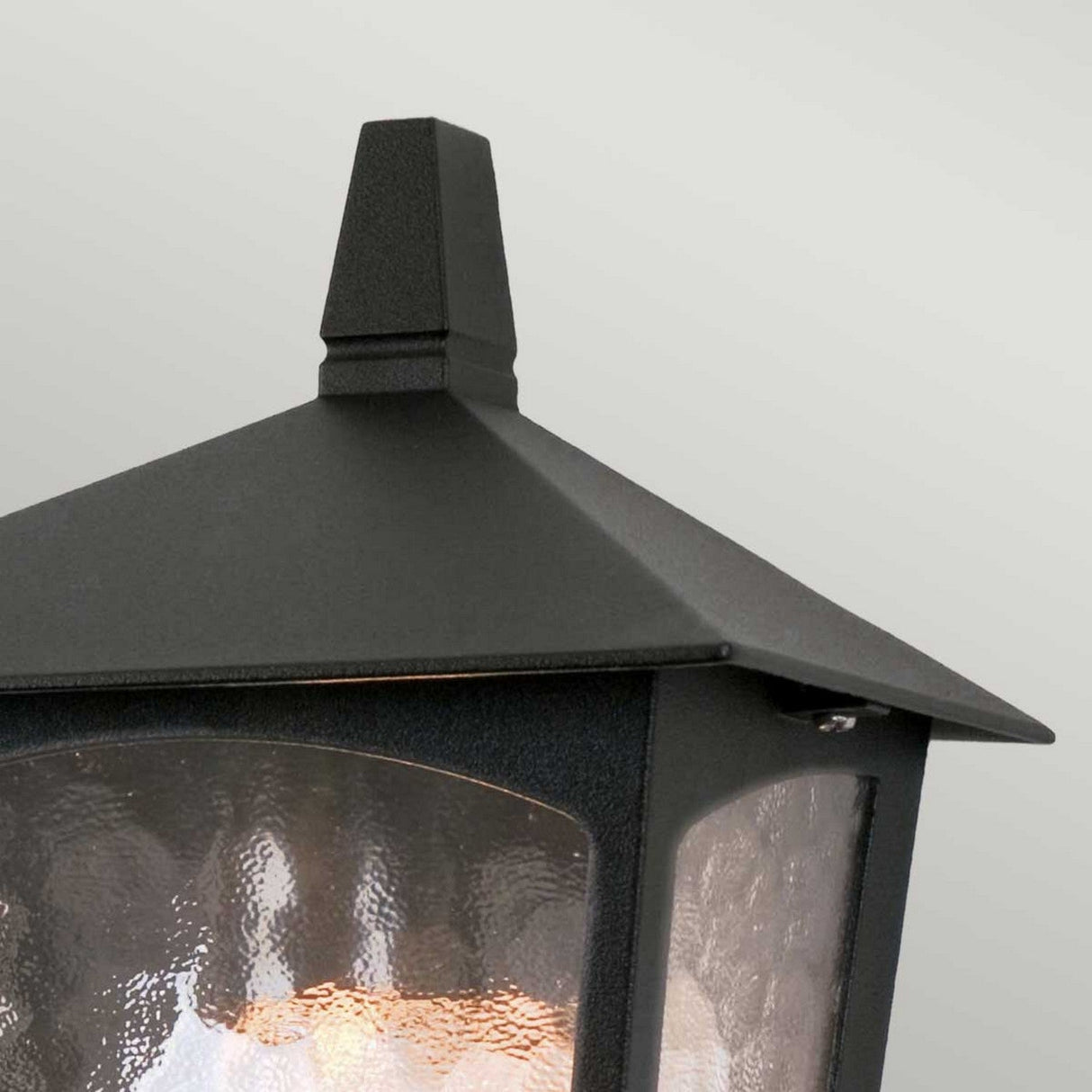 Close-up of the York 1 Light Outdoor Flush Light in black, featuring a die-cast aluminium lantern-style design with a triangular roof and glass panels. The softly glowing light inside highlights its traditional elegance against a plain, light gray background.