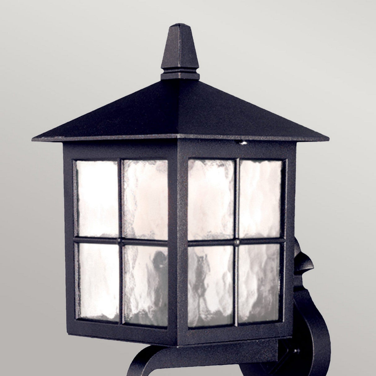 The Winchester 1 Light Outdoor Wall Up Light - Black is a wall lantern made from die-cast aluminum in black metal, showcasing frosted glass panels within its square design topped with a pointed finial. Its decorative mounting bracket adds a touch of historical elegance to the plain light gray background.
