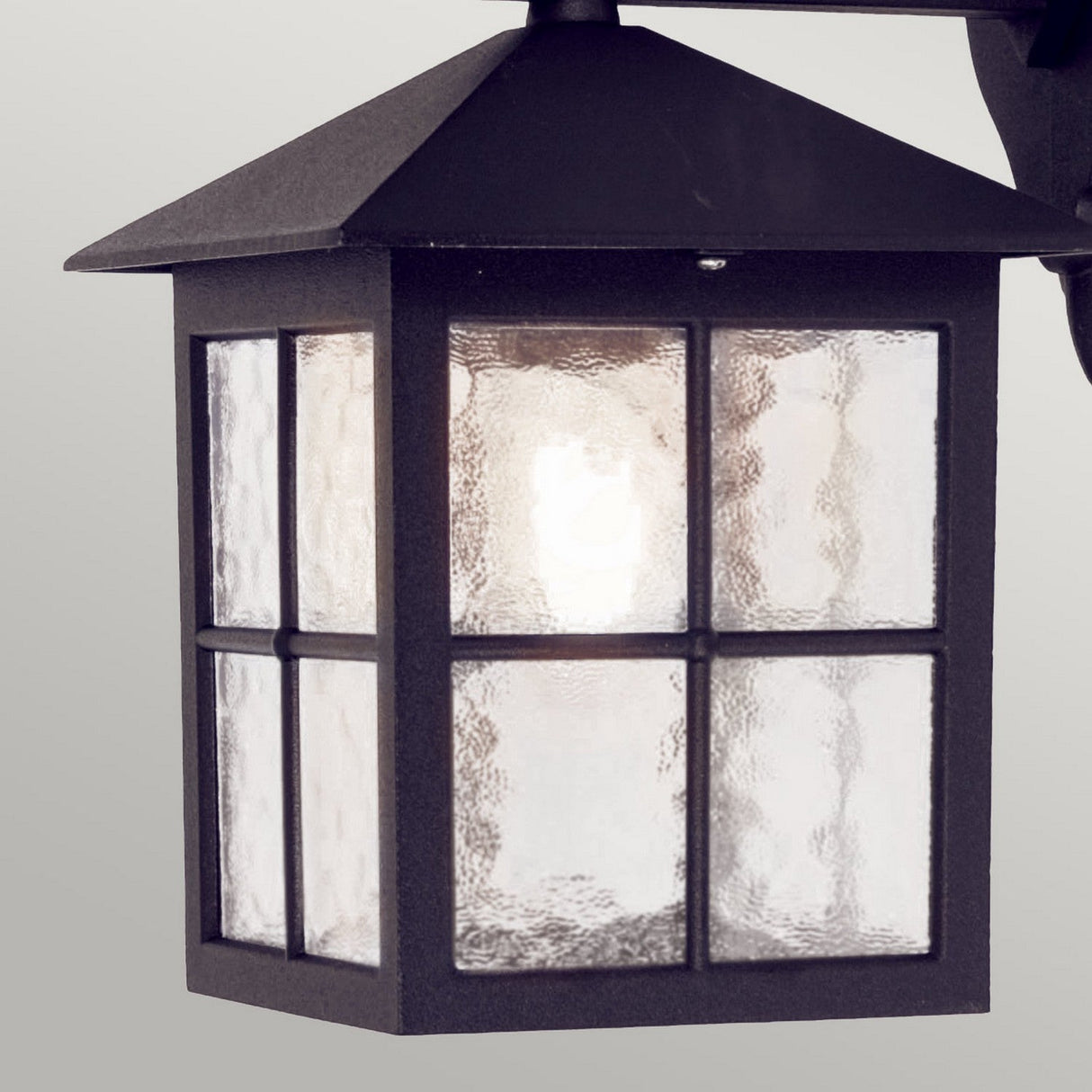 A close-up view of the Winchester-1 1 Light Outdoor Wall Down Light - Black reveals its vintage-inspired square design. The lantern, constructed from durable die-cast aluminum, features a clear, textured glass pane and is lit from within, highlighting its traditional geometric charm against a plain background.