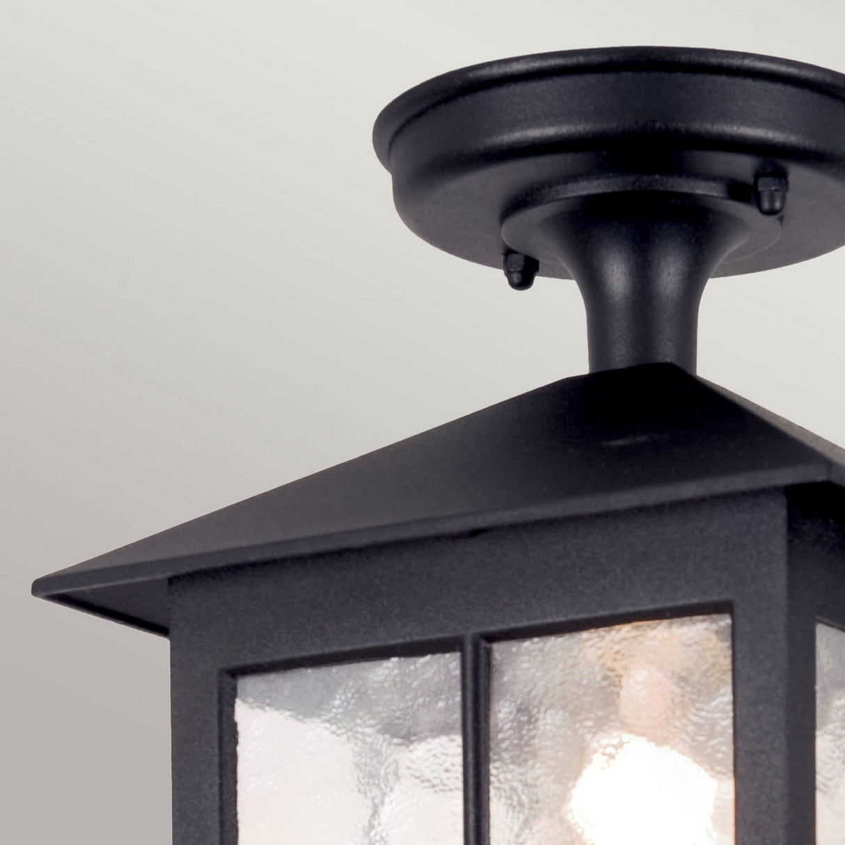 Close-up of the Winchester 1 Light Outdoor Porch Light - Black, a ceiling fixture for outdoor use featuring vintage charm. Its pyramid-shaped top and textured glass panels emit a warm glow, beautifully enhancing outdoor spaces against a neutral background.