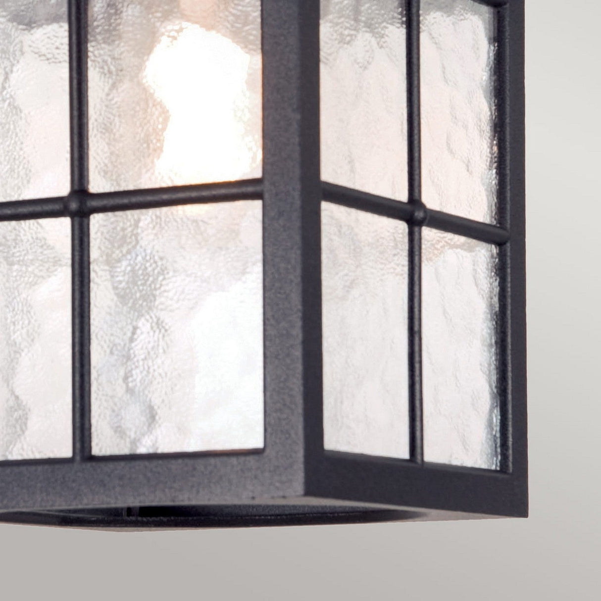A close-up view reveals a pendant light fixture with a black frame and textured glass, radiating vintage charm. The illuminated light casts a warm glow akin to the Winchester 1 Light Outdoor Porch Light - Black through its patterned glass panes, set against a neutral gray background.