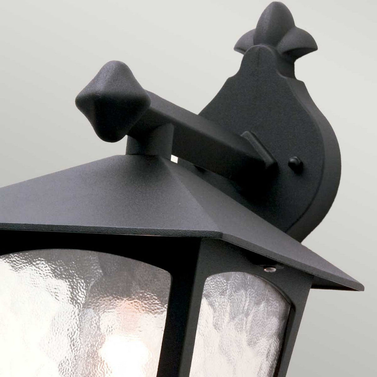 Close-up of the York 1 Light Outdoor Wall Down Light in black, showcasing a vintage design with a textured glass shade. The lamp features a decorative black metal frame embellished with a fleur-de-lis motif on top, providing charm and elegance to your garden décor.