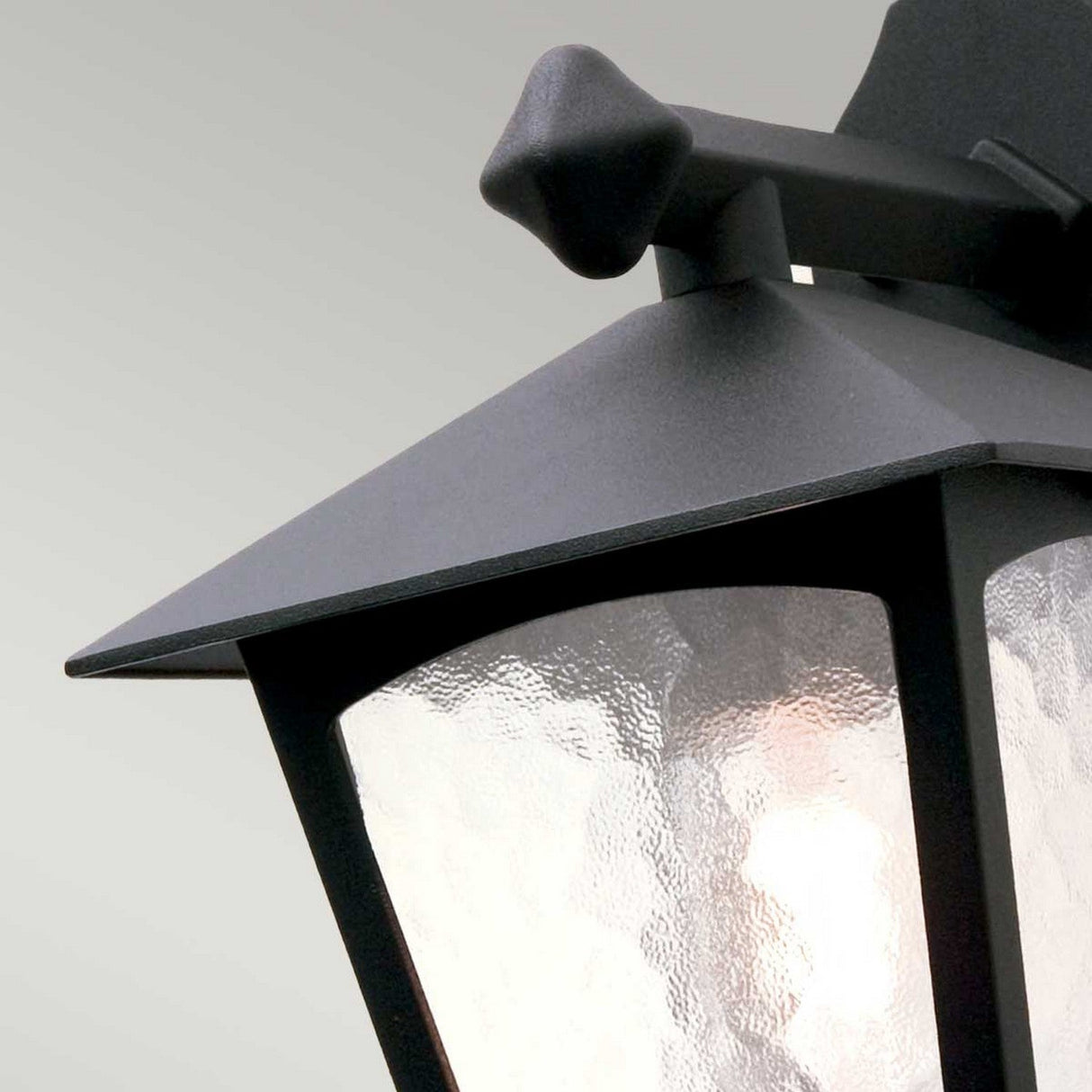 Close-up of the York 1 Light Outdoor Wall Down Light in black, boasting a modern design with a pointed top and textured glass panels. A soft glow emanates from this elegant garden wall light, casting warmth against a neutral background.