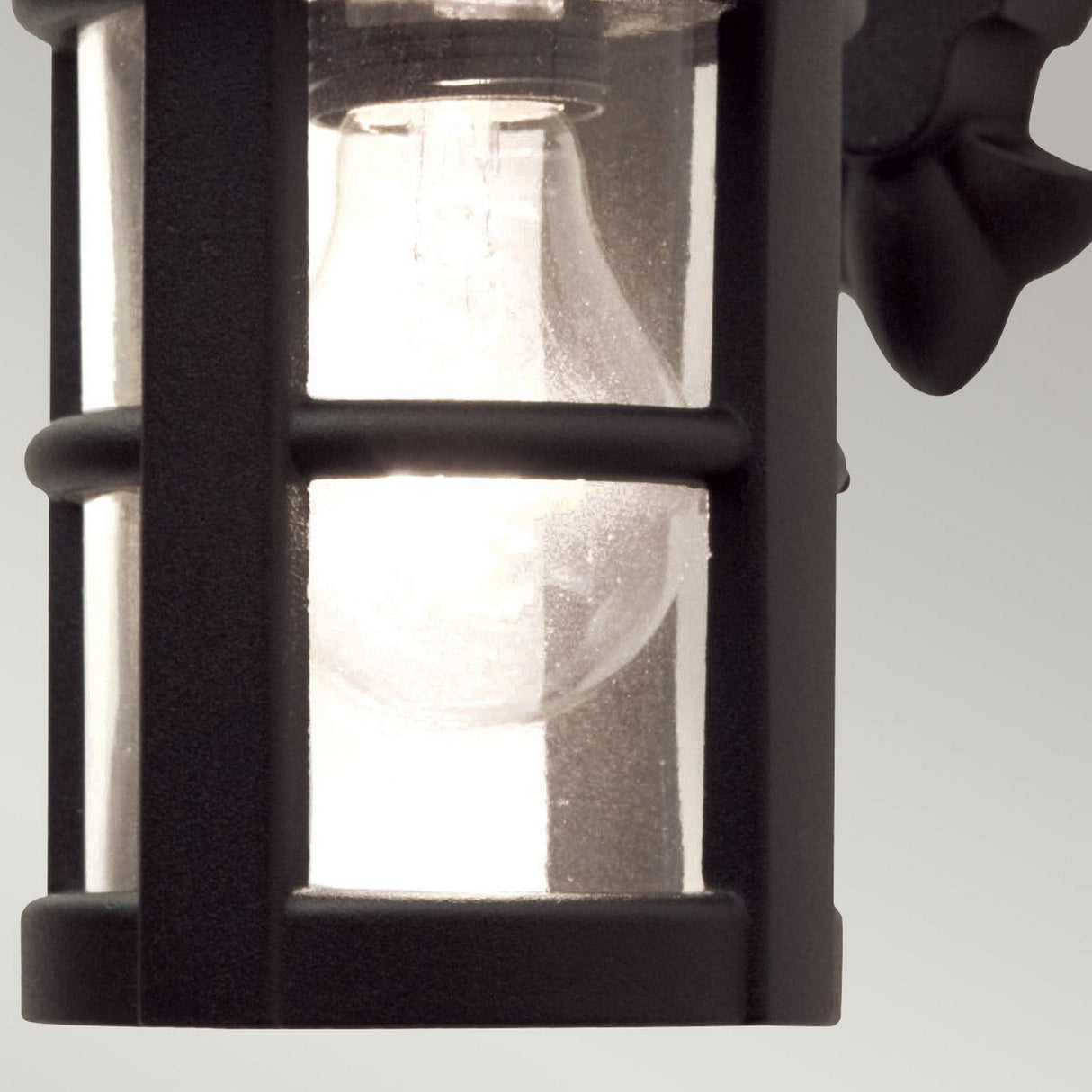 A close-up of a glowing bulb inside the Hereford 1 Light Outdoor Wall Down Lantern showcases its black, die-cast aluminum frame and glass grid, creating a rustic appearance. This large outdoor wall light stands out against the neutral gray background.