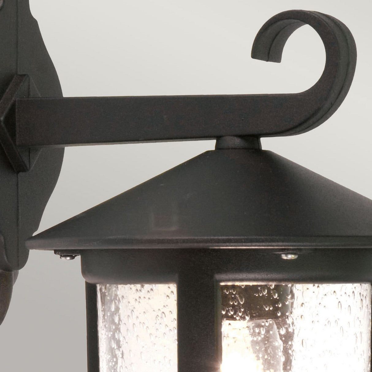 Close-up of the Hereford 1 Light Outdoor Large Wall Down Lantern - Black, featuring a classic lantern style with a curved arm and clear textured glass. The illuminated lantern highlights droplets on its glass surface, set against a neutral background.