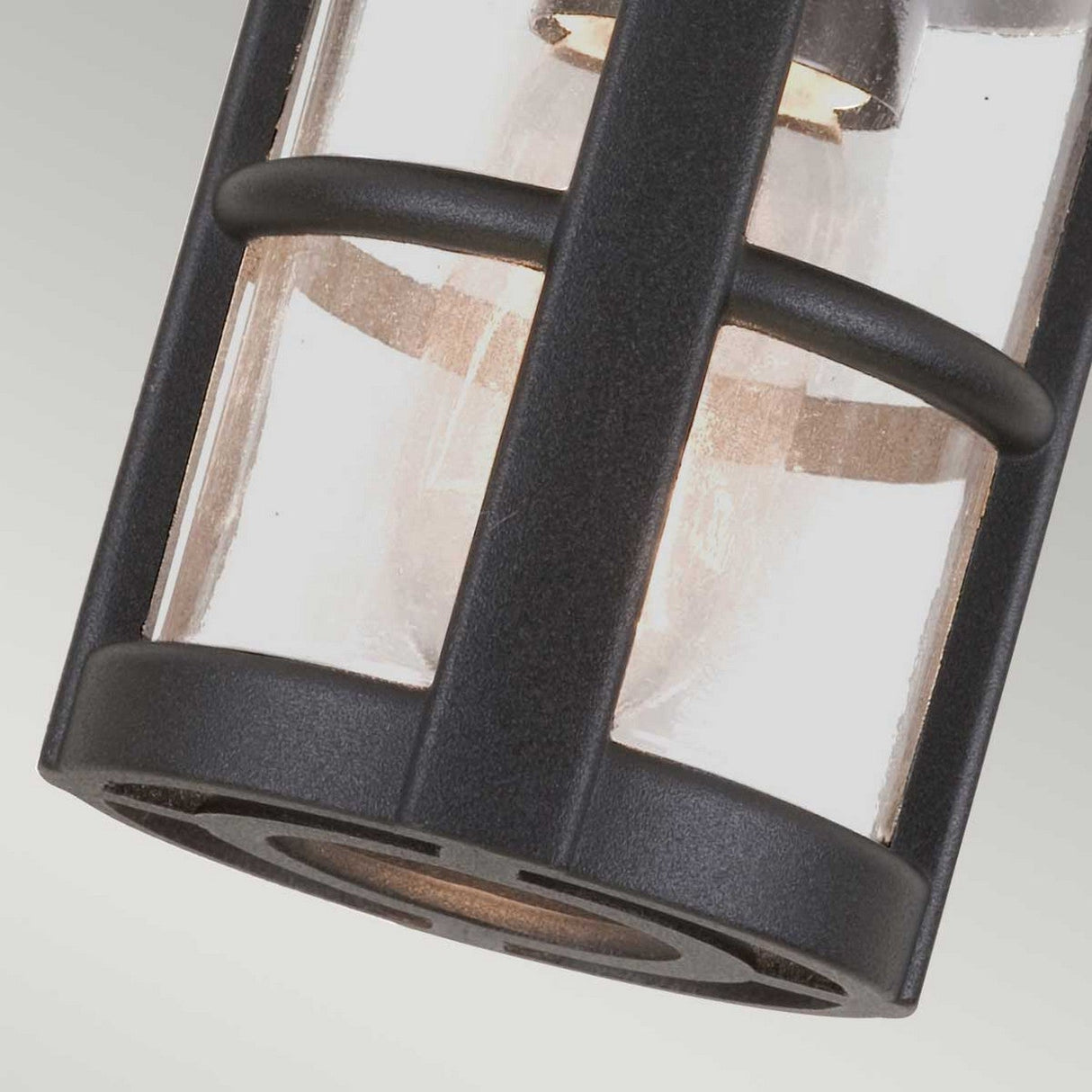 A close-up of the Hereford-2 1 Light Outdoor Porch Lantern - Black showcases its transparent cylindrical glass enclosure accentuated by vertical and horizontal black metal bars. Reminiscent of a traditional English lantern, the internal bulb softly illuminates this modern outdoor lighting fixture.