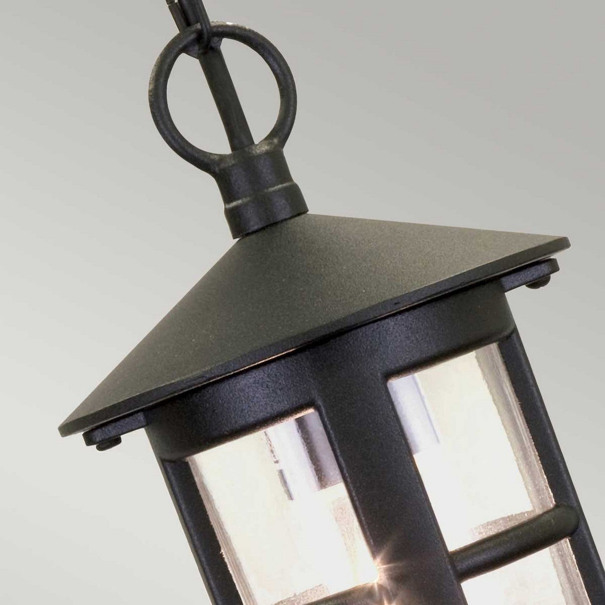 Here's a close-up of the Hereford 1 Light Outdoor Chain Pendant in Black, showcasing its classic lantern design with a pyramid-shaped top and transparent panels that reveal the bulb within. A circular ring is affixed at the top for easy hanging, making it an elegant outdoor lighting option.