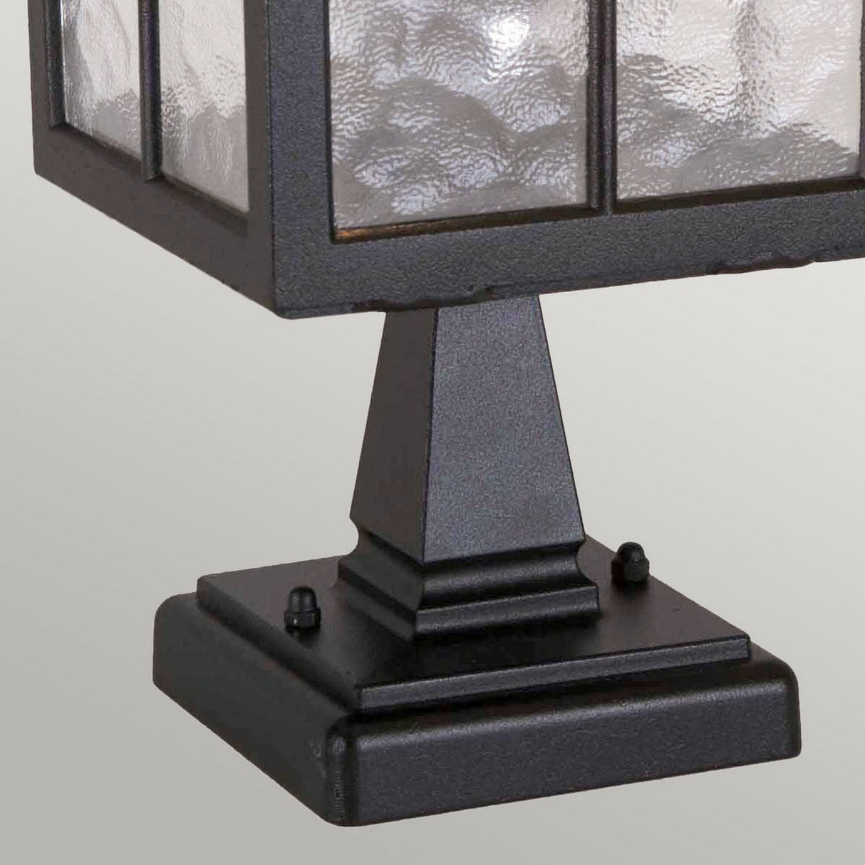 A close-up of the Winchester 1 Light Pedestal Light - Black showcases its die-cast aluminum construction and textured glass pane. Featuring a square base and rectangular frame, this outdoor lantern captures vintage charm against a plain, light gray background.
